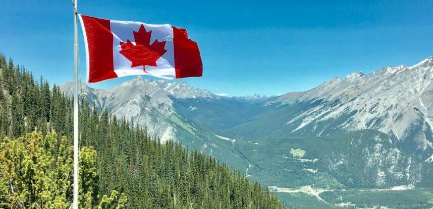 Canadian flag waving atop a scenic mountain view with blue skies and lush greenery.