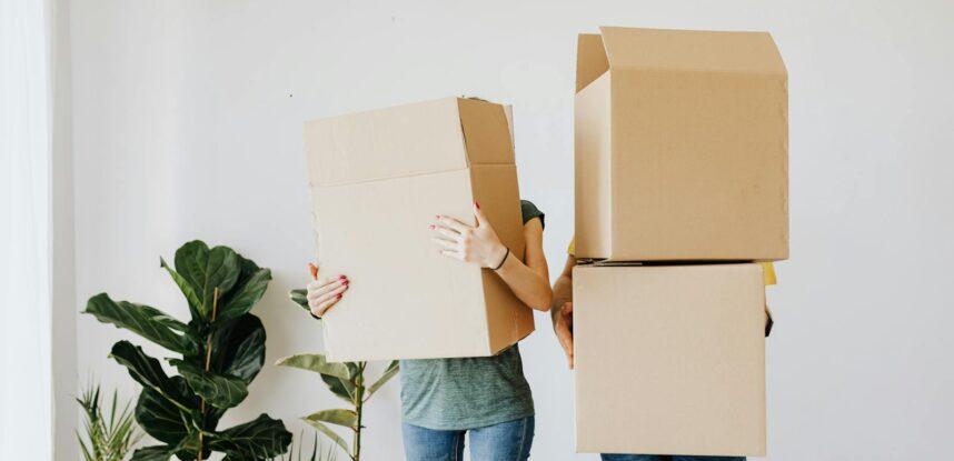 Unrecognizable couple wearing jeans standing carrying stacked carton boxes out of apartment during renovation on daytime